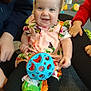 baby, child, smiling, toy, ball, floral_dress, indoor, carpet, happy, person, hand, play, colorful, legs, sitting, cute, infant, people, floor, casual