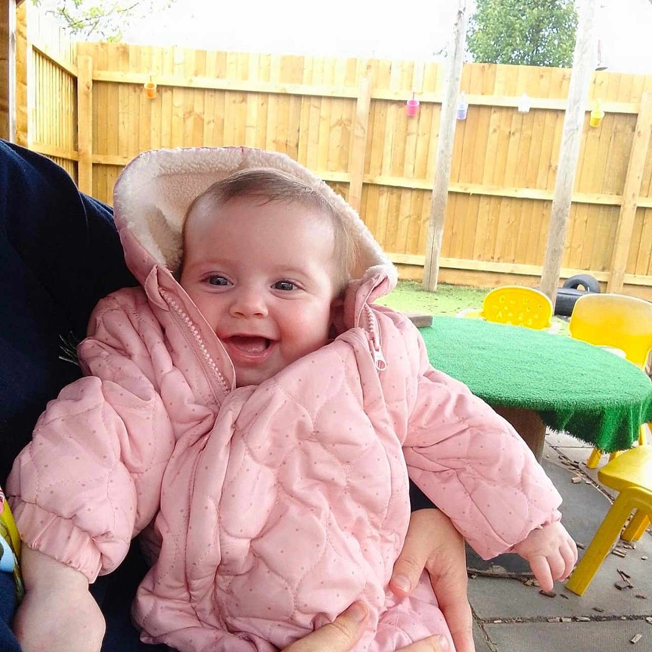 Skyla-Jayne is registered to the contest to win money with this photo: baby, chair, child, cold_weather, daylight, fence, green_table, hand, happy, holding, infant, jacket, outdoor, person, pink_jacket, play_area, smiling, table, wooden_fence, yellow_chair