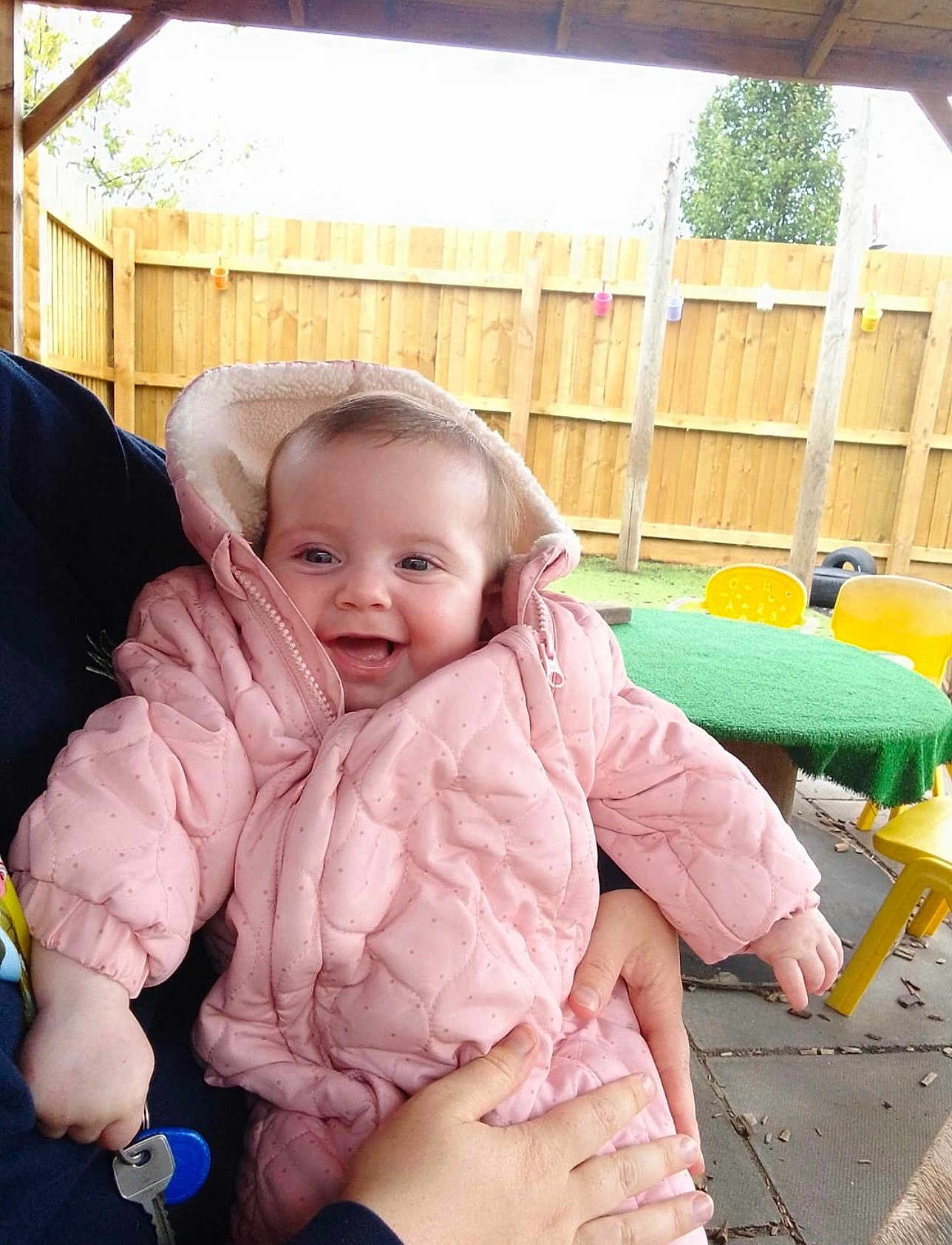 Skyla-Jayne is registered to the contest to win money with this photo: baby, pink_jacket, smiling, child, outdoor, fence, table, chair, hand, person, happy, play_area, cold_weather, jacket, holding, infant, wooden_fence, green_table, yellow_chair, daylight