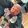 baby, infant, dinosaur_onesie, hand, sleeve, adult, living_room, wooden_floor, furniture, couch, chair, person, face, closeup, cozy, indoor, casual_clothing, skin, young_child, holding