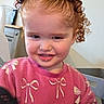 child, toddler, curly_hair, purple_scrunchies, pink_sweater, bows, smile, indoor, home_decor, cabinet, picture_frame, happy, cute, person, face, young_child, seated, furniture, cozy, portrait