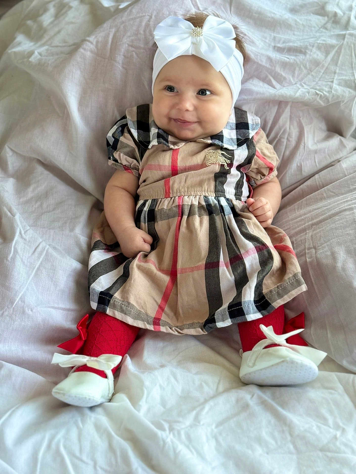 Delilah-Louise is registered to the contest to win money with this photo: baby, child, smiling, headband, bow, dress, plaid, red_tights, white_shoes, sitting, indoors, fabric, cute, infant, person, portrait, happy, playful, cozy, light