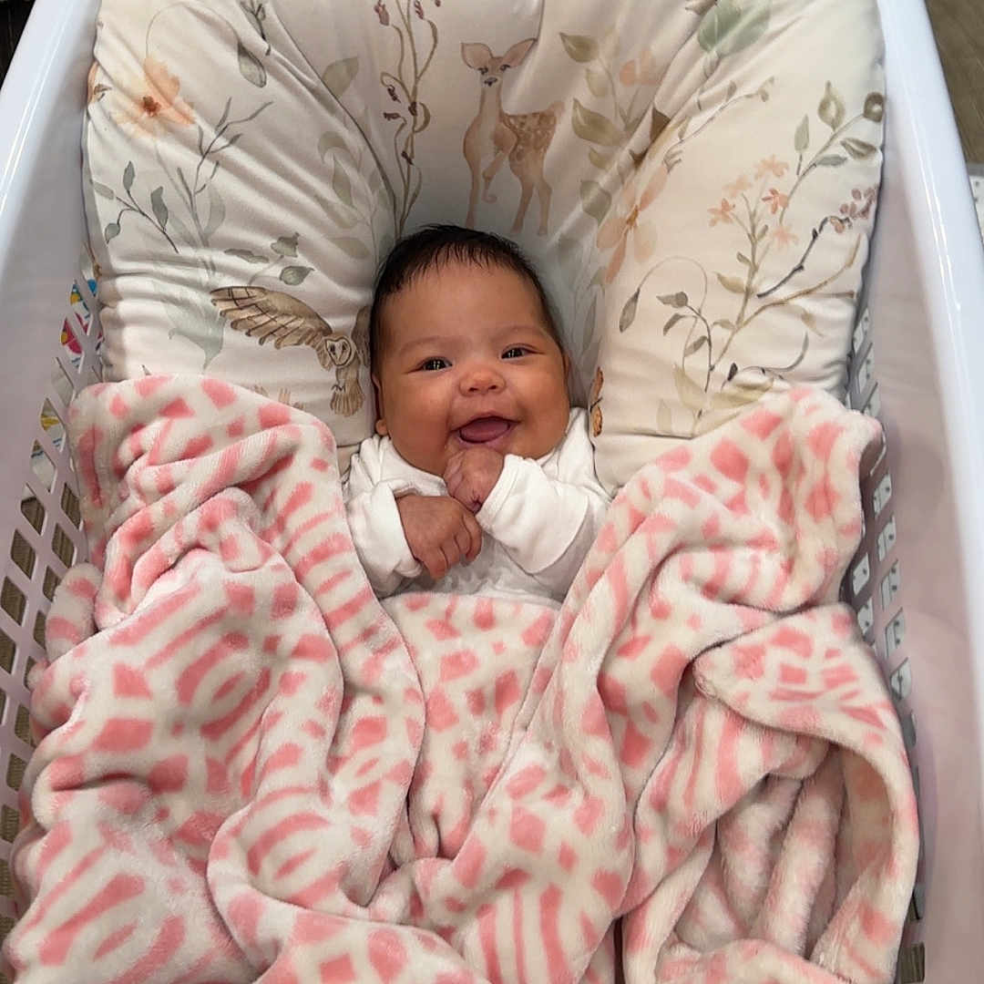 Zara is registered to the contest to win money with this photo: baby, blanket, child, comfort, cozy, cushion, cute, face, floral_pattern, happy, indoor, infant, laundry_basket, person, pink, portrait, resting, smiling, soft, white