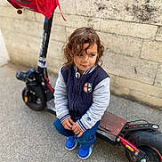 Josuè participe au concours pour gagner de l'argent avec cette photo : child, curly_hair, jacket, blue_sneakers, electric_scooter, backpack, stone_wall, outdoor, sidewalk, crouching, thoughtful, person, casual_clothing, urban, toy, transportation, young_child, street, footwear, daytime