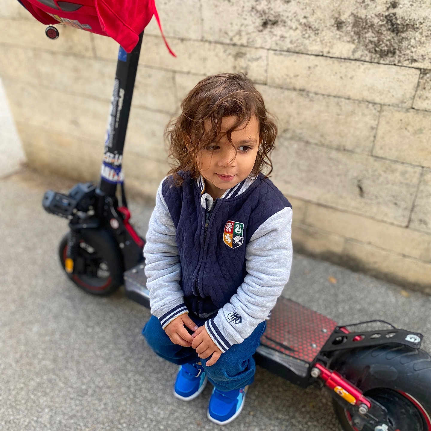 Josuè participe au concours pour gagner de l'argent avec cette photo : backpack, blue_sneakers, casual_clothing, child, crouching, curly_hair, daytime, electric_scooter, footwear, jacket, outdoor, person, sidewalk, stone_wall, street, thoughtful, toy, transportation, urban, young_child