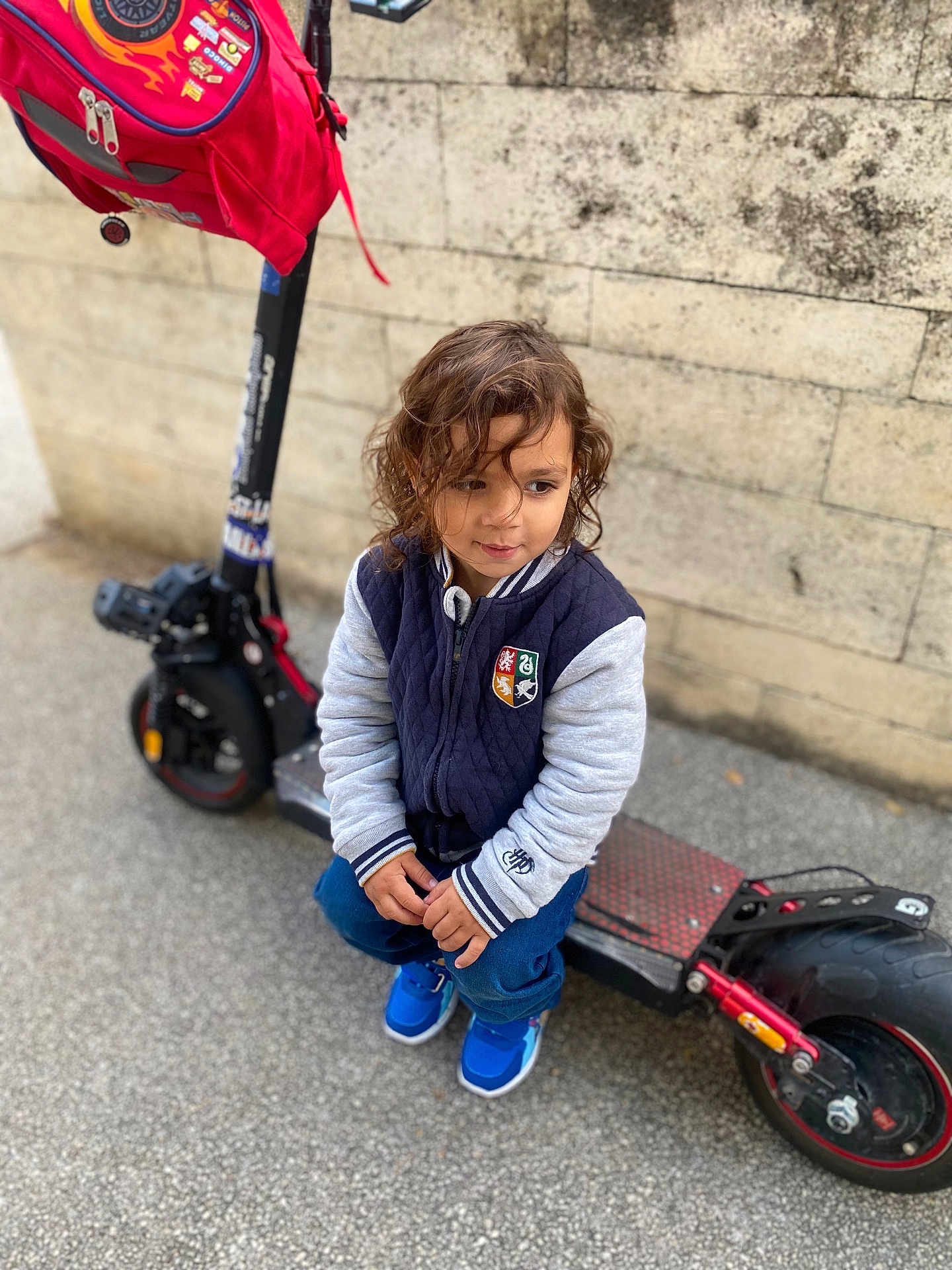 Josuè participe au concours pour gagner de l'argent avec cette photo : child, curly_hair, jacket, blue_sneakers, electric_scooter, backpack, stone_wall, outdoor, sidewalk, crouching, thoughtful, person, casual_clothing, urban, toy, transportation, young_child, street, footwear, daytime