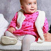 Lola a rejoint le concours — aidez-le/la à gagner de superbes lots ! baby, child, pink_dress, white_vest, gold_shoes, tights, sitting, plush_surface, portrait, indoor, curly_hair, cute, infant, looking_away, soft_lighting, neutral_background, hand, footwear, clothing, smiling