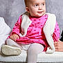 baby, child, pink_dress, white_vest, gold_shoes, tights, sitting, plush_surface, portrait, indoor, curly_hair, cute, infant, looking_away, soft_lighting, neutral_background, hand, footwear, clothing, smiling