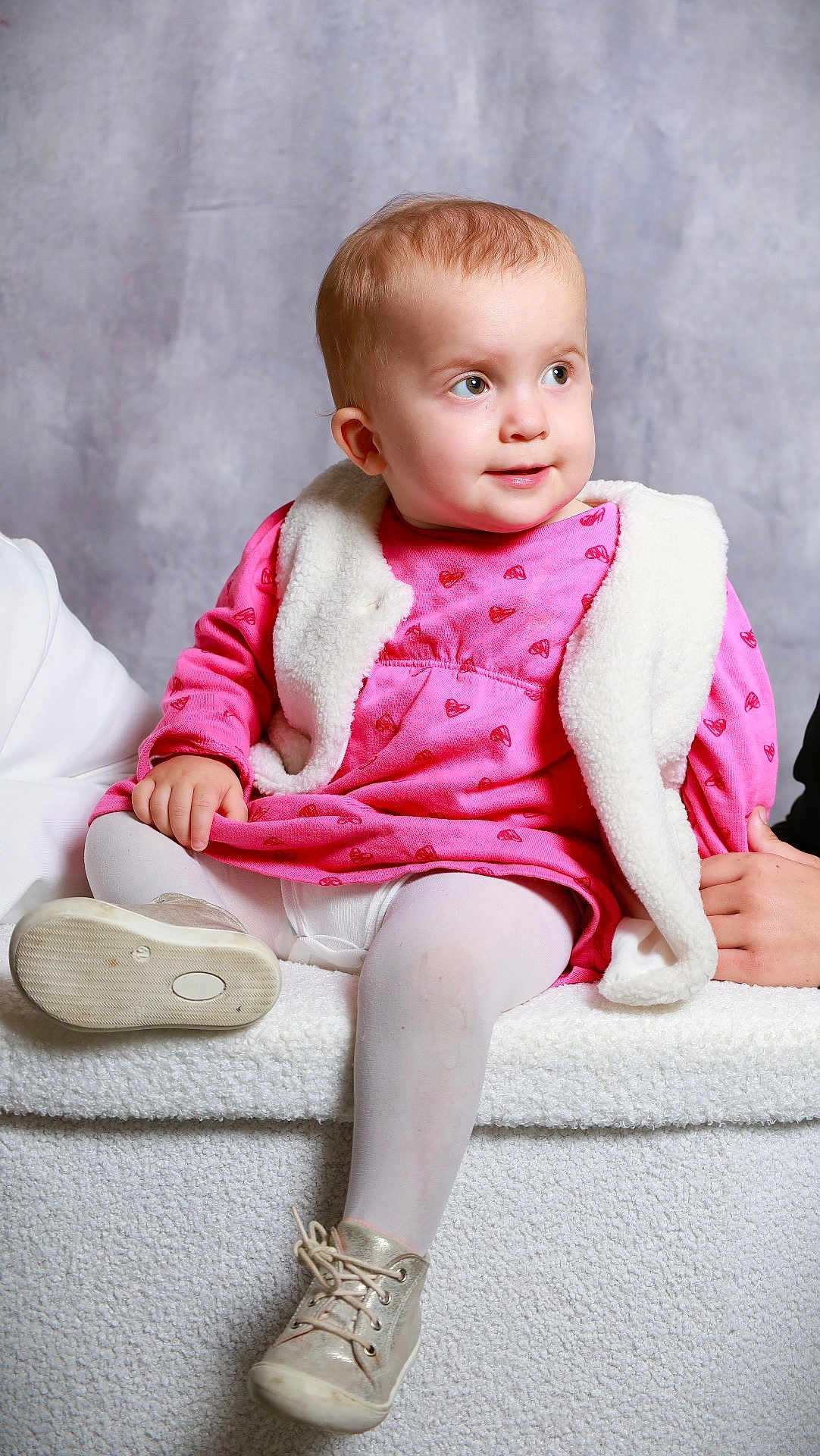Lola a rejoint le concours — aidez-le/la à gagner de superbes lots ! baby, child, pink_dress, white_vest, gold_shoes, tights, sitting, plush_surface, portrait, indoor, curly_hair, cute, infant, looking_away, soft_lighting, neutral_background, hand, footwear, clothing, smiling