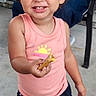 Adaline is registered to the contest to win money with this photo: toddler, child, smiling, happy, pink_tank_top, black_shorts, food, hand, outdoor, concrete_floor, person, curly_hair, joyful, casual_clothing, barefoot, sun_graphic, shorts, cheerful, portrait, daylight