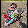child, tricycle, sunglasses, heart_shaped, outdoor, sunny, leafy_branch, smiling, casual_clothing, grass, tree_trunk, pavement, toy_vehicle, footwear, playful, young_kid, daylight, happy, person, recreational