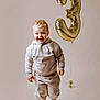 toddler, child, smiling, balloon, number_three, beige_clothing, hoodie, pants, striped_socks, studio, neutral_background, happy, walking, gold_balloon, celebration, joyful, portrait, indoor, cute, young_child