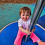 Madelyn is registered to the contest to win money with this photo: child, toddler, smiling, playground, blue_surface, pink_pants, sneakers, metal_pole, play_equipment, outdoor, rubber_play_surface, wood_chips, face, hands, portrait, happy, casual_clothing, eye_contact, small_child, play