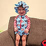 child, toddler, smile, hat, sunhat, swimsuit, american_flag_pattern, stripes, stars, red_white_blue, couch, sofa, living_room, bare_feet, standing, hands, face, portrait, toy, upholstery