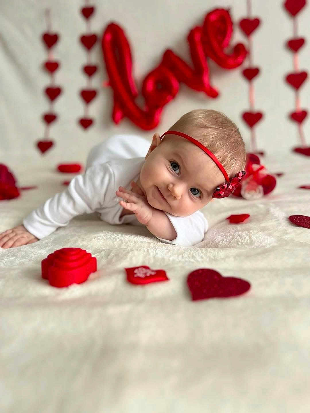 Arya is registered to the contest to win money with this photo: baby, infant, headband, red_bow, heart_decorations, love_balloon, white_blanket, lying_down, cute, portrait, holiday_theme, valentines_day, decorations, close_up, indoor, child, adorable, smiling, soft_texture, celebration
