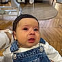 baby, child, denim_overalls, white_shirt, tears, crying, indoor, wooden_floor, person_holding, face, emotion, closeup, young_child, infant, candid, human, clothing, hand, furniture, home