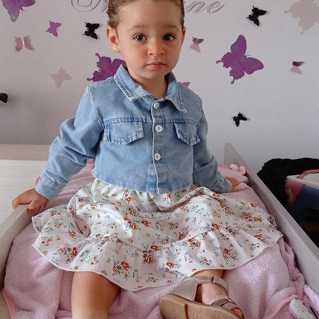 Maélane a rejoint le concours — aidez-le/la à gagner de superbes lots ! bed, butterfly_wall_decals, child, clothing, cute, denim_jacket, earrings, feet, floral_skirt, furniture, girl, hair_tie, indoor, pink_blanket, portrait, room_decor, sandals, serious_expression, skin, toddler