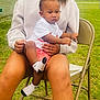toddler, child, adult, sitting, outdoor, grass, chair, white_shirt, red_shorts, socks, folding_chair, holding, person, casual_clothing, greenery, daylight, brick_wall, curly_hair, relaxed, family