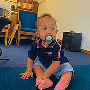 Charvis Jr joined the competition — help win amazing prizes! baby, pacifier, child, indoor, blue_cushion, striped_shorts, navy_shirt, wooden_chair, speaker, carpet, curly_hair, sitting, barefoot, cute, cross_legged, furniture, person, looking_away, floor, room