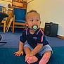 baby, pacifier, child, indoor, blue_cushion, striped_shorts, navy_shirt, wooden_chair, speaker, carpet, curly_hair, sitting, barefoot, cute, cross_legged, furniture, person, looking_away, floor, room