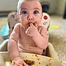baby, child, high_chair, messy, food, face, hand, blue_eyes, curious, indoor, sitting, eating, cute, infant, skin, fingers, tray, blurred_background, expression, closeup