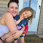 Felicity is registered to the contest to win money with this photo: woman, child, hug, outdoor, door, welcome_mat, smile, curly_hair, sneakers, casual_clothing, concrete, front_porch, home, person, family, affection, door_handle, window, blue_shirt, pink_shoes
