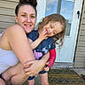 woman, child, hug, outdoor, door, welcome_mat, smile, curly_hair, sneakers, casual_clothing, concrete, front_porch, home, person, family, affection, door_handle, window, blue_shirt, pink_shoes