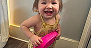 Lilyana joined the competition — help win amazing prizes! toddler, child, smiling, yellow_outfit, barefoot, indoor, wooden_floor, pink_toy, happy, standing, face, person, water_jug, floor, hair, playful, expression, home, curtain, radiator