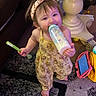 Lilyana joined the competition — help win amazing prizes! toddler, child, baby_bottle, spoon, headband, yellow_outfit, barefoot, carpet, tablet, toy, table_leg, indoor, person, smiling, standing, playful, cute, home, floor, furniture