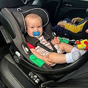 Noah is registered to the contest to win money with this photo: baby, pacifier, car_seat, car_interior, child, seat_belt, toy, clothing, window, person, socks, book, seat, headrest, black_leather, safety, infant, child_reading, vehicle, family