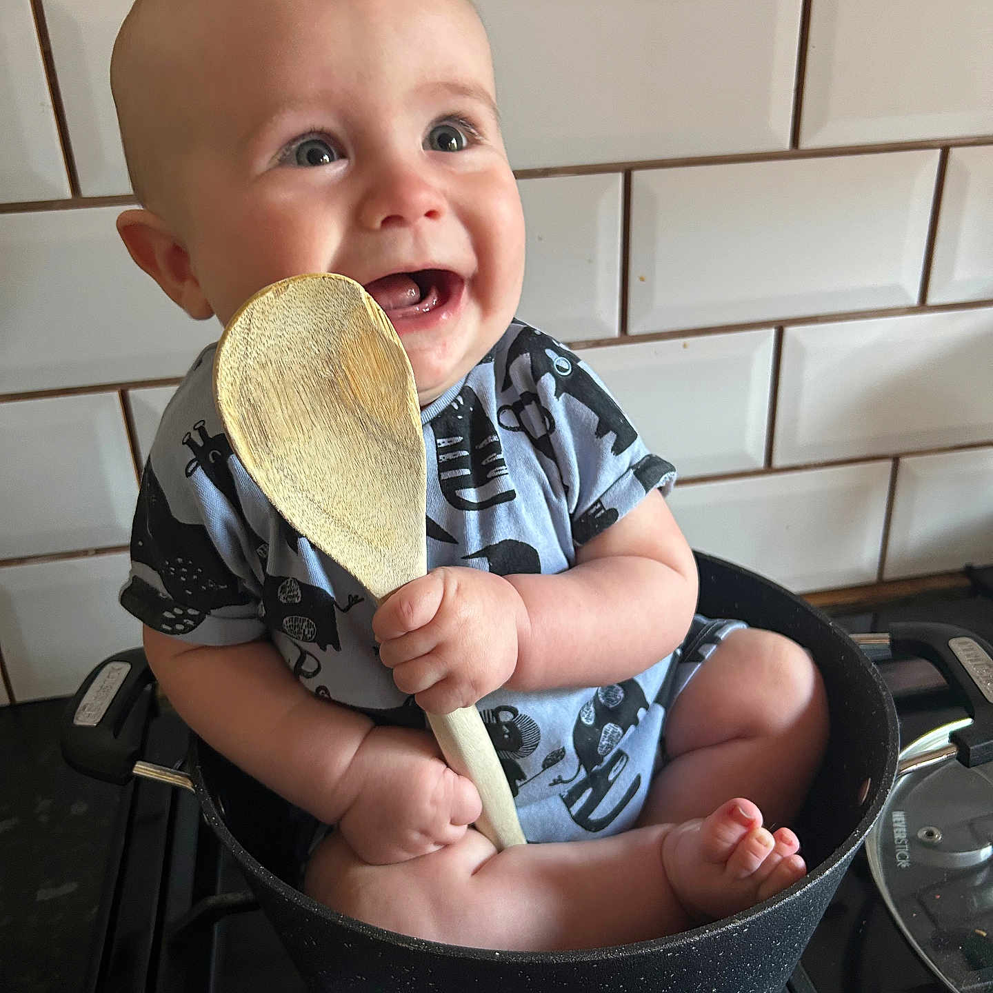 Noah is registered to the contest to win money with this photo: baby, barefoot, child, clothing, cooking_utensil, cute, happy, holding, indoors, infant, kitchen, kitchenware, person, playful, pot, sitting, smile, stove, tile_wall, wooden_spoon