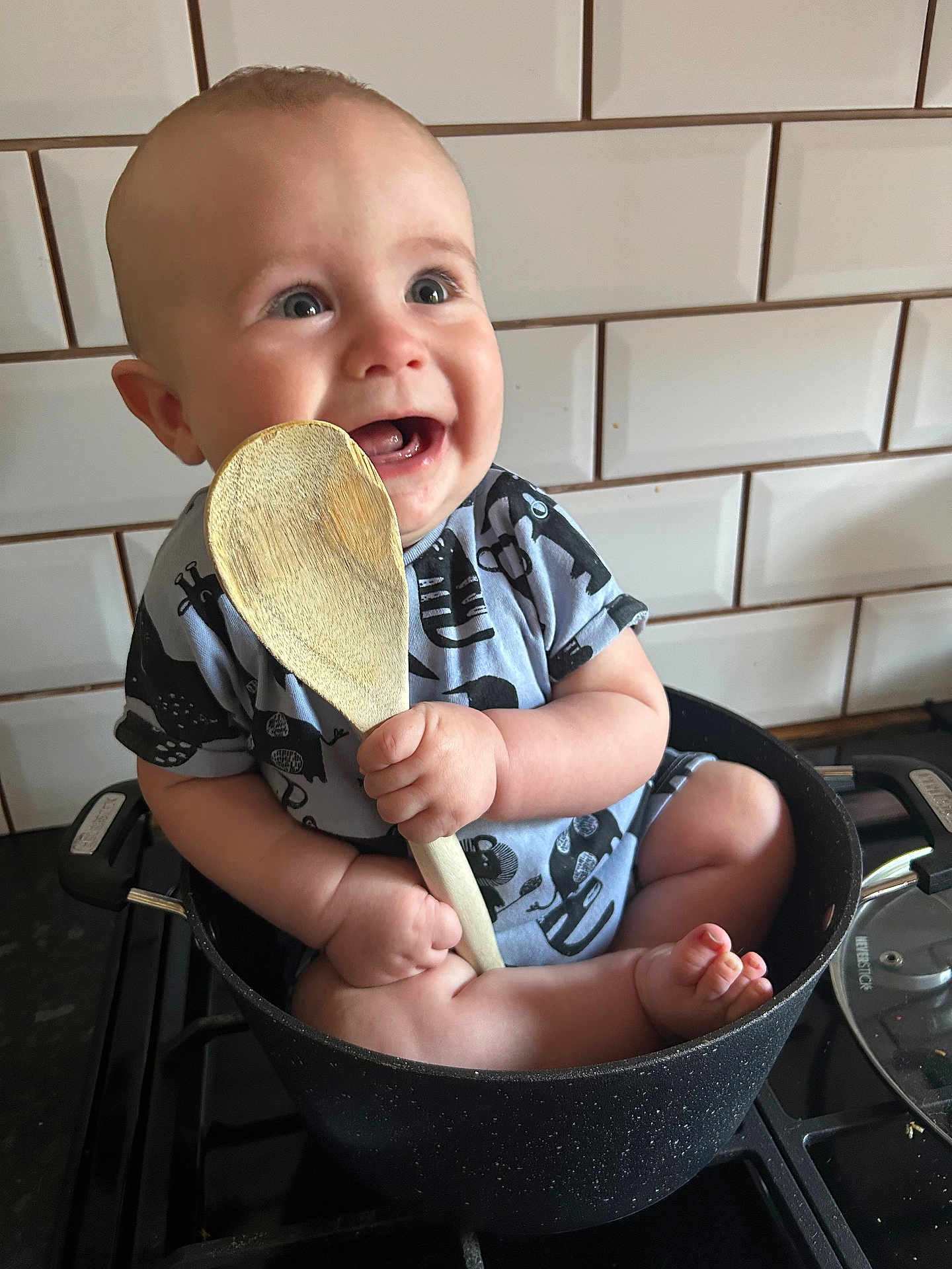 Noah is registered to the contest to win money with this photo: baby, child, pot, kitchen, stove, wooden_spoon, smile, happy, sitting, tile_wall, infant, cute, cooking_utensil, kitchenware, person, indoors, playful, clothing, barefoot, holding