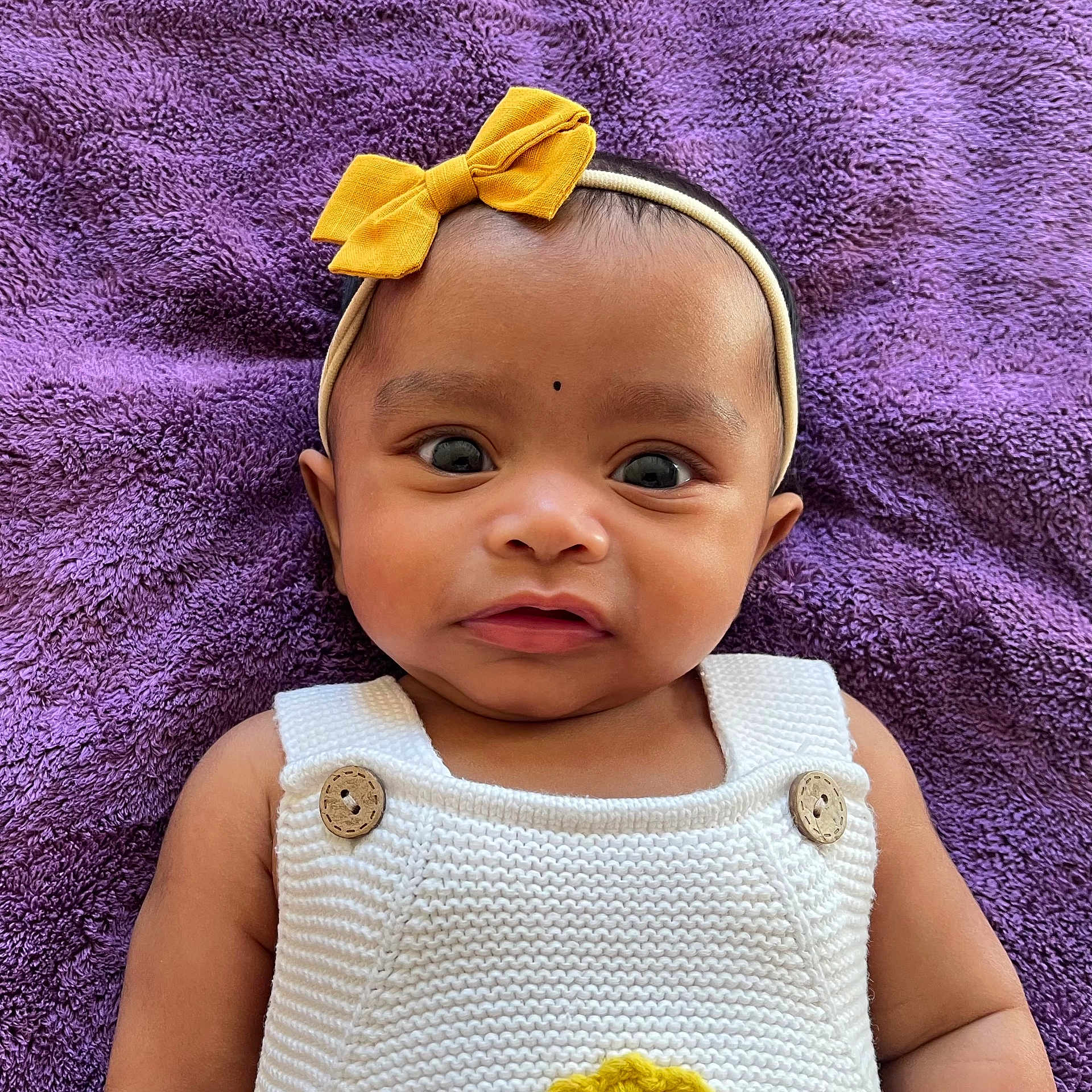 Gnazhini is registered to the contest to win money with this photo: baby, infant, face, headband, bow, purple_blanket, knitted_outfit, white_clothing, wooden_buttons, curious_expression, lying_down, closeup, child, cute, person, portrait, skin, eyes, mouth, indoors
