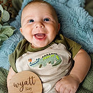 Wyatt joined the competition — help win amazing prizes! baby, smiling, blue_eyes, infant, blanket, soft_texture, embroidered_clothing, crocodile_design, wooden_plaque, name_tag, cute, portrait, cozy, happy, child, person, indoor, closeup, skin, hand