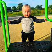 Luke joined the competition — help win amazing prizes! toddler, child, playground, play_equipment, green_bars, outdoor, sunny, blue_sky, grass, trees, young_child, casual_clothing, black_pants, gray_shirt, holding, standing, daylight, serious_expression, person, recreation
