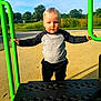 toddler, child, playground, play_equipment, green_bars, outdoor, sunny, blue_sky, grass, trees, young_child, casual_clothing, black_pants, gray_shirt, holding, standing, daylight, serious_expression, person, recreation