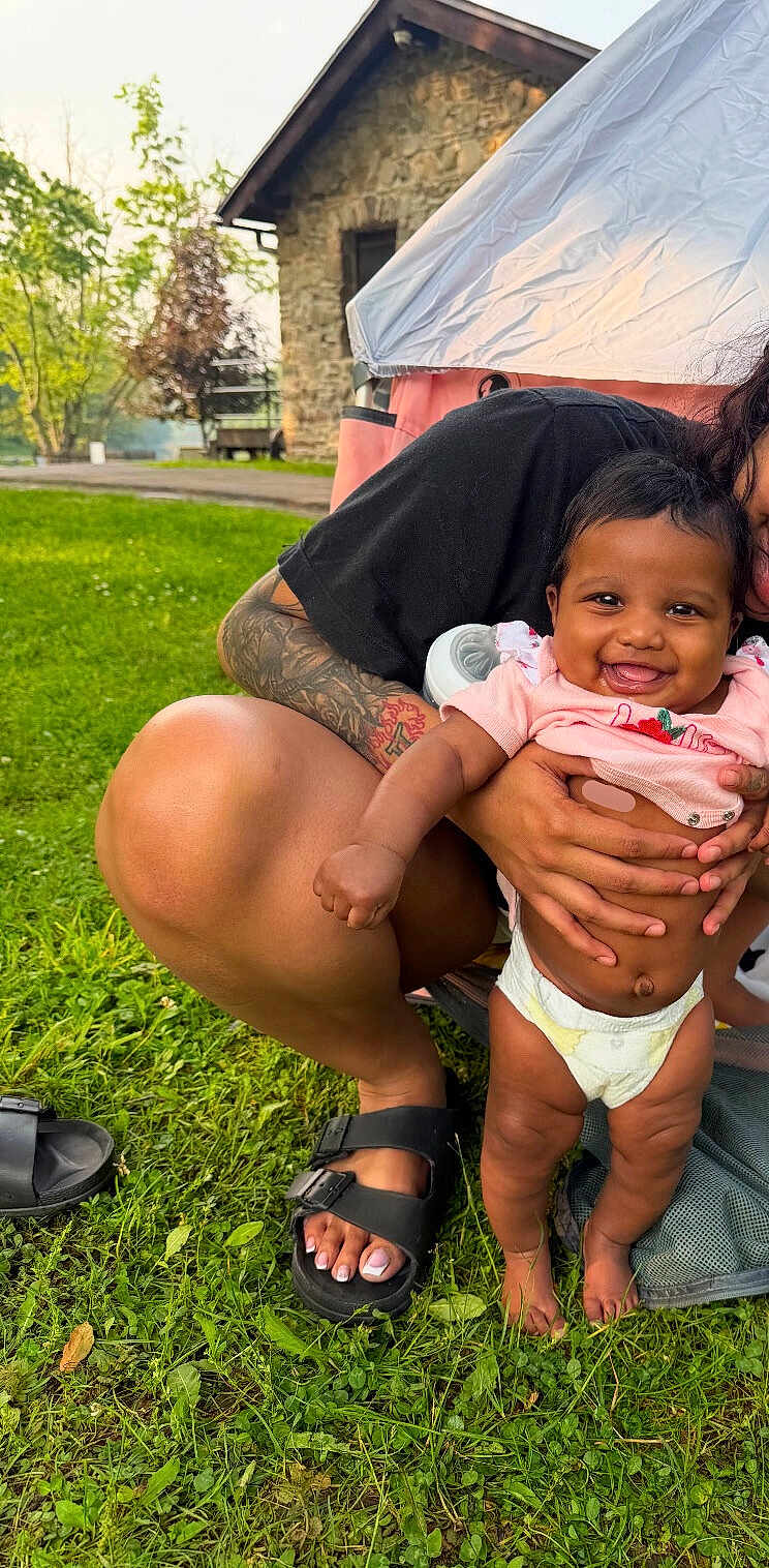 Zenaaliyah is registered to the contest to win money with this photo: baby, smiling, infant, child, diaper, pink_clothing, tattoo, adult, grass, outdoor, greenery, stone_building, sandals, person, holding, happy, nature, sunlight, crouching, hand