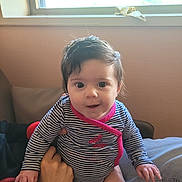 Octavia a rejoint le concours — aidez-le/la à gagner de superbes lots ! baby, child, striped_clothing, pink_trim, hand, indoor, window, natural_light, curly_hair, sitting, person, face, cute, infant, portrait, looking_at_camera, furniture, home, soft_light, expression