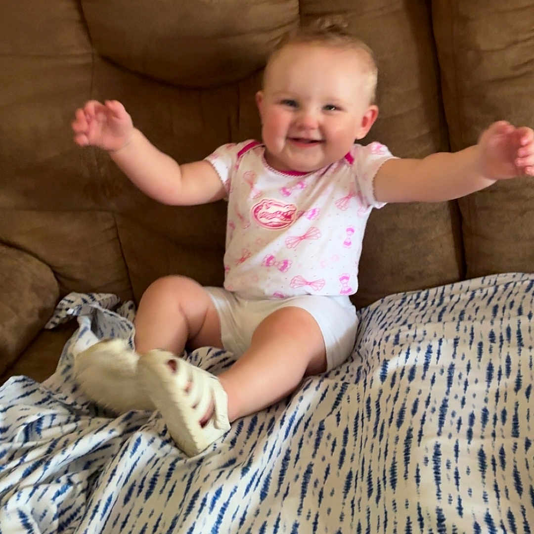 Wrenlee is registered to the contest to win money with this photo: baby, child, smiling, sitting, couch, blanket, indoor, happy, person, footwear, white_clothing, pink_pattern, face, arms, legs, soft_shoes, home, comfort, infant, cute