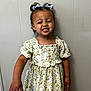 child, girl, smiling, floral_dress, bows, hair_accessories, happy, portrait, standing, indoor, casual_clothing, cute, young, skin_tone, person, expression, childhood, fashion, wall, natural_light