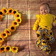Victoire a rejoint le concours — aidez-le/la à gagner de superbes lots ! adorable, baby, baby_fashion, bright_colors, child, cute, dimples, floral_pattern, happy, indoors, infant, lying_down, number_two, patterned_pants, playful, portrait, smiling, sunflowers, wooden_floor, yellow_clothing
