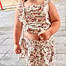 child, girl, curly_hair, pigtails, floral_outfit, top, skirt, shoes, standing, pavement, outdoor, portrait, cute, young_child, fashion, daylight, expression, person, casual_clothing, street