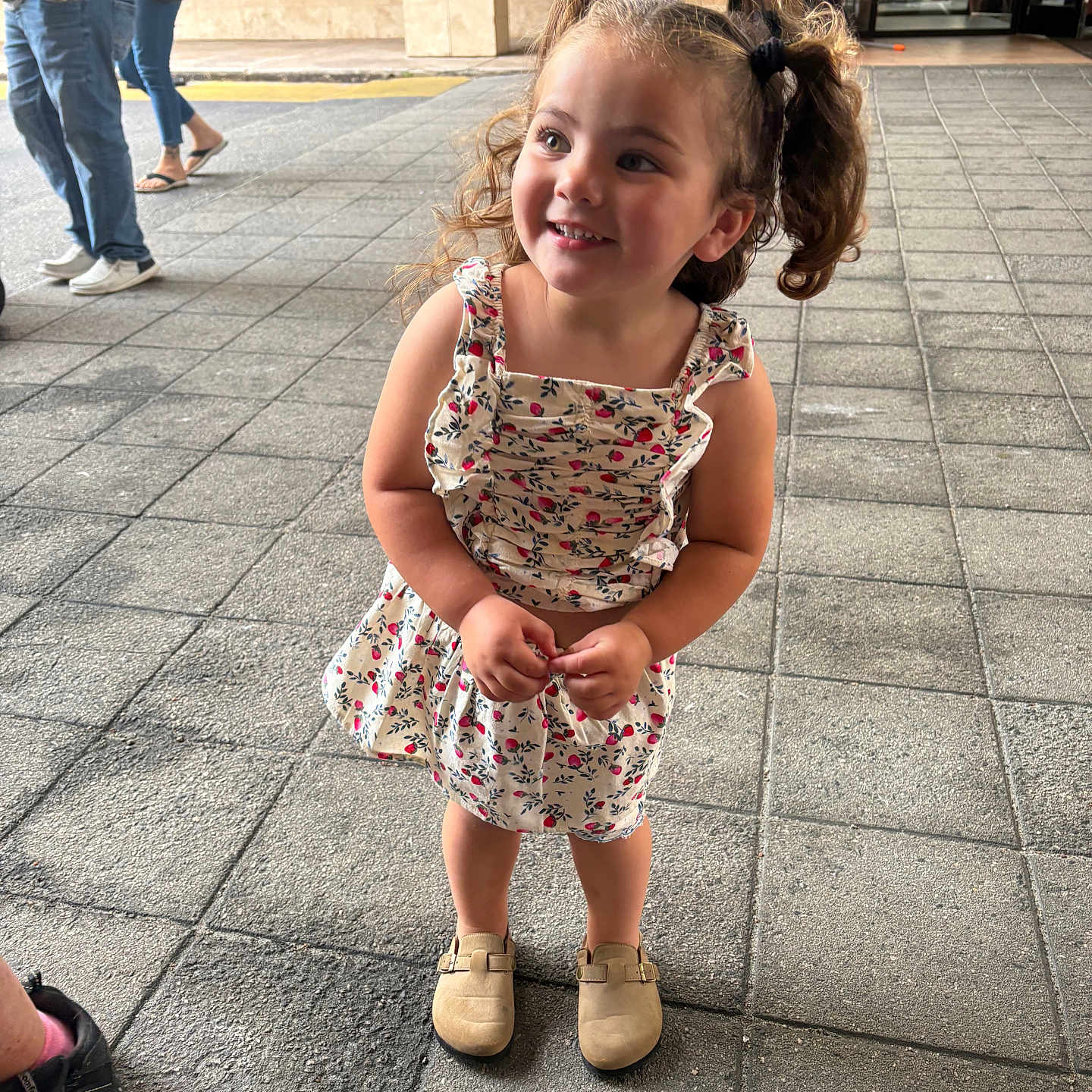 Kaylee is registered to the contest to win money with this photo: child, girl, smiling, curly_hair, pigtails, floral_dress, beige_shoes, sidewalk, tiles, outdoor, people, walking, building, entrance, casual_clothing, happy, portrait, daylight, standing, person