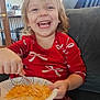toddler, child, smiling, red_dress, bow_pattern, paper_plate, fork, shredded_cheese, couch, living_room, window, natural_light, happy, eating, indoor, person, furniture, blond_hair, casual_clothing, home