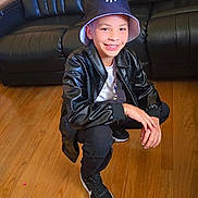 Yosbel joined the competition — help win amazing prizes! child, young_boy, smiling, portrait, crouching, indoor, living_room, sofa, hardwood_floor, bucket_hat, baseball_cap, leather_jacket, black_clothing, sneakers, fashion, casual_style, person, face, furniture, photography