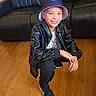 child, young_boy, smiling, portrait, crouching, indoor, living_room, sofa, hardwood_floor, bucket_hat, baseball_cap, leather_jacket, black_clothing, sneakers, fashion, casual_style, person, face, furniture, photography