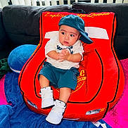 Noa Daniel is registered to the contest to win money with this photo: baby, child, cap, sneakers, red_chair, car_shaped_chair, couch, blanket, blue_blanket, pink_blanket, clothing, indoor, person, seated, hands, looking_away, footwear, casual_wear, cute, plush