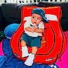 baby, child, cap, sneakers, red_chair, car_shaped_chair, couch, blanket, blue_blanket, pink_blanket, clothing, indoor, person, seated, hands, looking_away, footwear, casual_wear, cute, plush
