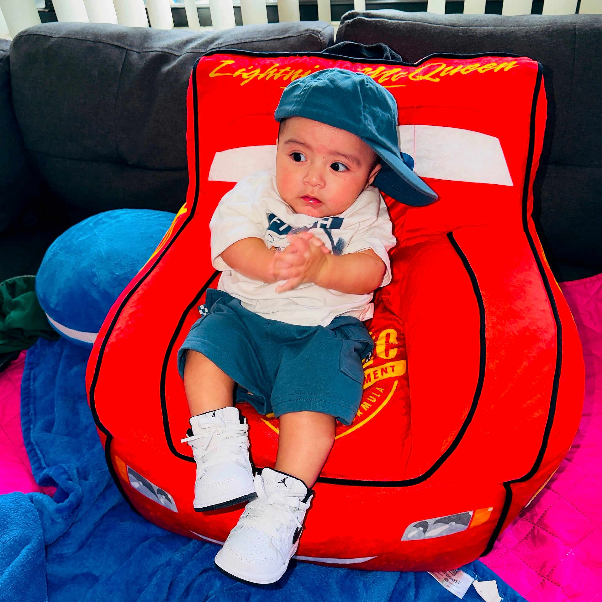 Noa Daniel is registered to the contest to win money with this photo: baby, child, cap, sneakers, red_chair, car_shaped_chair, couch, blanket, blue_blanket, pink_blanket, clothing, indoor, person, seated, hands, looking_away, footwear, casual_wear, cute, plush