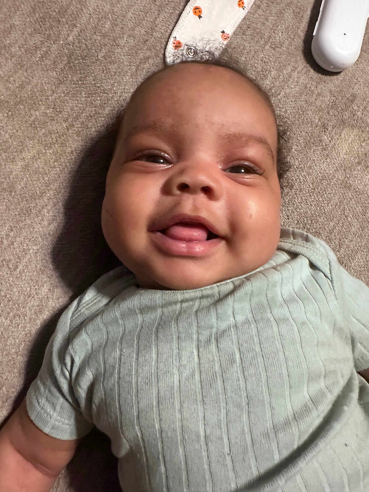 Shai joined the competition — help win amazing prizes! baby, infant, smiling, face, tongue, carpet, clothing, ribbed_shirt, lying_down, happy, cute, child, skin, head, expression, indoor, toy, fabric, closeup, person